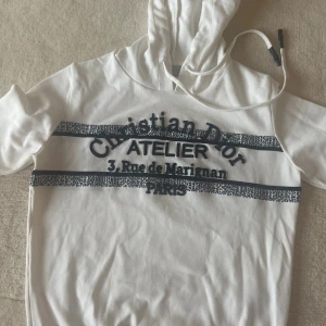 Vit hoodie  - Säljer en vit hoodie med svart broderad text och grafiska detaljer framtill. Tröjan har huva med snörning Snygg 