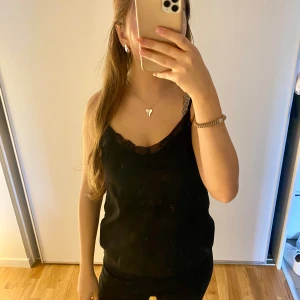 Svart linnetopp med spetskant - Snygg svart linnetopp med tunna axelband och v-ringning dekorerad med spetskant. Perfekt att styla med jeans eller kjol för en trendig look. Toppens material känns mjukt och följsamt mot huden.