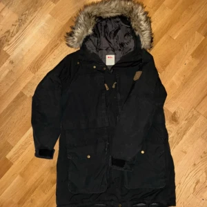 Svart Barents Parka från Fjällräven - Tidlös och varm parka för vinterdagar både i stan och under enklare friluftsäventyr. Parkan har ett membran som håller vind och väta ute, en värmande syntetfyllning och flera praktiska fickor. Parkan ger härlig värme över stussen och den stora huvan har fleecetyg på insidan och stänger ute kylan. Syntetpälsen går att ta av om så önskas. Jackan erbjuder bra förvaring i de stora praktiska fickorna, samt en tvåvägsdragkedja.  Material: Återvunnen Polyamid och Polyester