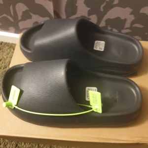 Adidas Yeezy Slide svart strl 40.5 - Svarta Adidas Yeezy slides Tillverkade i syntetmaterial för en clean look och bekväm känsla. Perfekta för att matcha med streetwear. Kommer med originalbox och tagg.