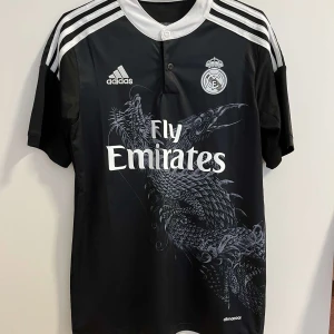 Real Madrid Ronaldo customtröja - Säljer en svart Real Madrid fotbollströja från Adidas. Tröjan har ett stort drakmotiv på framsidan, vita detaljer och tryck, samt Ronaldo 7 på ryggen. Tillverkad i lätt och ventilerande climacool-material. Storlek S.