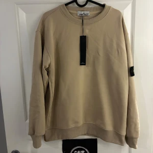 Beige sweatshirt från Stone Island - Säljer en exklusiv helt ny och oanvänd beige sweatshirt från Stone Island i storlek Small i en oversize modell med klassisk patch på vänster ärm. Tröjan har rund halsringning, ribbade muddar och är tillverkad i mjuk bomull. Perfekt för dig som gillar stilrena och trendiga plagg med streetkänsla.
