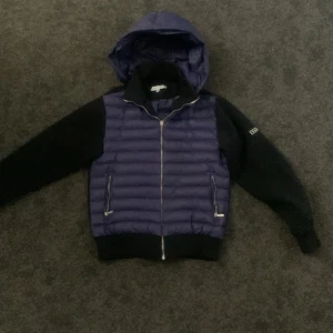 Blå och svart pufferjacka från LeVon - Snygg pufferjacka från LeVon i blått och svart med quiltad framsida och huva. Jackan har dragkedja framtill, två fickor med dragkedja och ribbade muddar. Ärmarna är svarta och har LeVon-logga på vänster ärm. Perfekt för kyliga dagar.