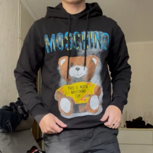 Svart Moschino Medium hoodie  - Den här tröjan är en svart hoodie från Moschino, en del av deras välkända “Teddy Bear” kollektion. Ikoniskt björntryck framtill och blått Moschino-tryck över bröstet. Tröjan har huva med snörning och långa ärmar. Tillverkad i 100% mjuk bomull för en skön och avslappnad look.