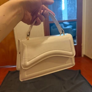 Vit handväska med axelrem - Stilren vit handväska i skinnimitation med snygg vågig detalj på locket. Väskan har ett kort handtag och en avtagbar axelrem med silverfärgat spänne. Perfekt storlek för det viktigaste och enkel att matcha till olika outfits.