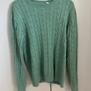 Grön kabelstickad tröja från GANT - Snygg grön kabelstickad tröja från GANT med rund halsringning och diskret logga på bröstet. I strl M
