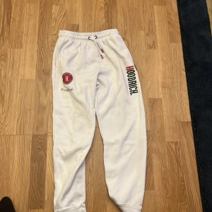 Vita mjukisbyxor från Hoodrich - Snygga vita mjukisbyxor från Hoodrich med tryck i svart och rött på benet och bak. Byxorna har elastisk midja med snörning och muddar vid bensluten. Perfekta för en chill och avslappnad stil.