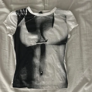 Vit och svart t-shirt med grafiskt tryck -  vit t-shirt med svart grafiskt tryck på framsidan som ger en edgy vibe. T-shirten har rund hals och korta ärmar, och är figurnära i modellen. Materialet känns som mjuk bomull, perfekt för dig som gillar statement-plagg.