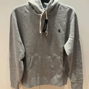 Grå hoodie från Polo Ralph Lauren M - Helt ny grå hoodie från Polo Ralph Lauren i storlek M, alla lappar kvar.Endast provad och passar tyvärr inte. Klassisk design med huva och snörning, stor magficka och broderad marinblå logga på bröstet. Nypris 2295kr