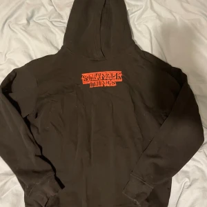 Stranger Things hoodie från H&M - Svart hoodie från H&M med Stranger Things-tryck i rött framtill och stor färgglad print på ryggen med karaktärer från serien. Klassisk huva och långa ärmar, perfekt för dig som älskar serien och vill sticka ut med en snygg streetstyle.