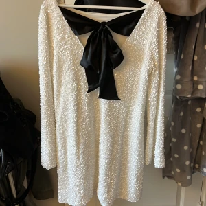 Vit paljettklänning med svart rosett - Supersnygg vit kortklänning från vero Moda täckt med glittriga paljetter och en stor svart rosett i satin baktill. Endast använd en gång. Klänningen har lång ärm och en djup v-ringning i ryggen. Perfekt för dig som vill sticka ut med en elegant och trendig look 🤍🖤