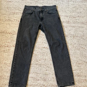 Svarta jeans från Nudie Jeans, W31 L30 - Snygga svarta jeans från Nudie Jeans med klassisk femficksdesign och raka ben. Jeansen har subtila sömmar på bakfickorna och är tillverkade i slitstarkt bomullstyg. Perfekt passform med normal midja och stilren look. Grity Jackson 