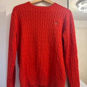 Röd kabelstickad tröja från GANT - Kabelstickad röd tröja från GANT med rund halsringning och broderad logga på bröstet. Tillverkad i 100% bomull och har ribbade muddar vid ärmslut och nederkant. Använd TVÅ gånger, precis som en helt ny tröja. Storlek 170cm (15-16 år men såklart kan den passa äldre eller yngre).