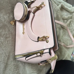 Ljusrosa handväska med gulddetaljer - Supersöt ljusrosa handväska från Bershka med guldfärgade detaljer och dragkedjor. Väskan har både handtag och axelrem samt en liten rund miniväska som hänger framtill. Tillverkad i syntetmaterial och har svart innerfoder. Perfekt för dig som gillar stilrena accessoarer.lite fläckar som syns på bild 1