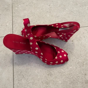 Nine West röda prickiga klackar  - Snygga röda wedges från Nine West med beige prickigt mönster. Skorna har öppen tå, kilklack och rem runt hälen. Tillverkade i satinliknande material som ger en glansig finish. Perfekta för dig som vill sticka ut med färg och mönster.