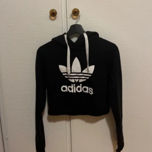 Svart croppad hoodie från Adidas - Svart croppad hoodie från Adidas med stor vit logga och text framtill. Tröjan har långa ärmar, huva med vita snören och en avslappnad passform. Perfekt för en sportig och trendig look. Aldrig använd 