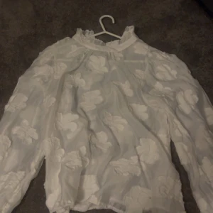 Vit blommig blus från H&M - Säljer en superfin vit blus från H&M i storlek 34. Blusen har transparent tyg med stora blommiga detaljer och långa ärmar. Den har en hög hals med rynk och är perfekt för dig som gillar romantisk och trendig stil.