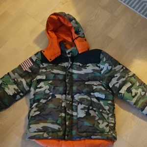 Camouflage pufferjacka från Ralph Lauren - Fet pufferjacka från Ralph Lauren Denim & Supply med camomönster och orange foder. Jackan har huva, stora fickor och amerikansk flag patch på ärmen. Perfekt för kalla dagar när du vill sticka ut. Oversized vibe och riktigt street.