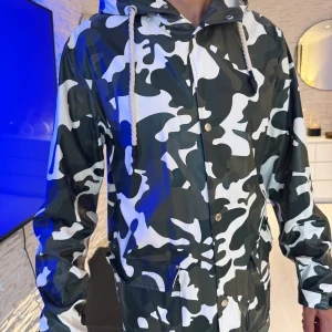 Camouflage regnjacka med huva - Snygg regnjacka med svart, grå och vit camouflage-mönster. Jackan har huva med dragsko, tryckknappar framtill och stora fickor. Perfekt för regniga dagar och streetstyle-vibe. Passar perfekt som oversize modell.