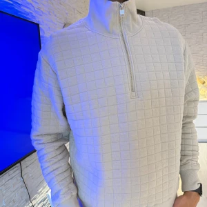 Beige quiltad half zip tröja - Säljer en beige långärmad tröja med quiltat rutmönster och hög krage med half zip-dragkedja. Tröjan har en relaxed passform och är tillverkad i mjukt material, perfekt för kyligare dagar. Enkel och stilren design som passar till det mesta. Passar perfekt som oversize modell.