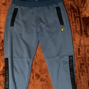 Blå mjukisbyxor från Lyle & Scott - Snygga blå mjukisbyxor från Lyle & Scott med svarta detaljer längs fickorna och logga i gult på ena benet. Byxorna har resår i midjan och svarta band med Lyle & Scott-tryck längs benen. Perfekta för chill eller träning.