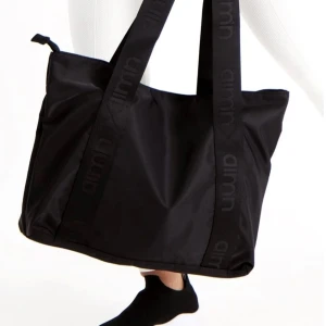 Svart axelväska från Aim'n - Stor svart axelväska från Aim'n med breda axelband där loggan är invävd. Väskan har dragkedja upptill och är tillverkad i slitstark polyester, perfekt för att bära med sig allt man behöver till träningen eller vardagen. Nypris 600kr.