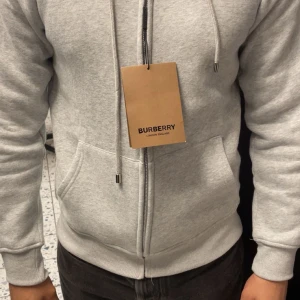 Grå hoodie från Burberry med dragkedja - Snygg ljusgrå hoodie från Burberry med klassiskt rutigt foder i huvan. Tröjan har dragkedja framtill, snörning i huvan och två stora fickor. Tillverkad i mjuk bomullsmix som känns riktigt skön mot huden. Perfekt för dig som gillar stilrena och exklusiva plagg. Helt ny och jätte bekväm känns lite mindre än M
