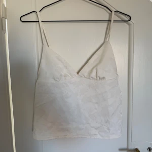Vit linnetopp i satin - Säljer en ljusvit linnetopp med tunna axelband och v-ringning. Toppen har ett subtilt blommigt mönster i tyget och är tillverkad i glansig satin som ger en lyxig känsla. Perfekt till jeans eller kjol för en fräsch look. Toppen är aldrig använd heller!🥰