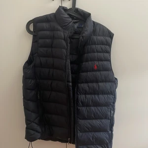 Marinblå dunväst från Polo Ralph Lauren - Nypris: 3599. I nyskick! Snygg marinblå dunväst från Polo Ralph Lauren med quiltad design och klassisk ståkrage. Västen har dragkedja framtill, två fickor med dragkedja och den ikoniska röda loggan broderad på bröstet. Perfekt för lager-på-lager och stilren look. Passar både S och M