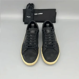 Svarta Saint Laurent sneakers - Svarta sneakers från Saint Laurent med handskriven logga på sidan. Skorna har svart canvas och detaljer i skinn, samt beige platt gummisula. Klassisk låg modell med snörning och rund tå. Kommer med originalpåse och extra skosnören.