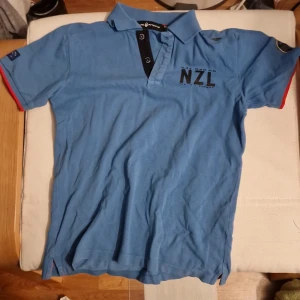 Sail Racing blå pikétröja, storlek 140 - Blå sail racing t-shirt från NZL Racing med svart knappslå och tryck på bröstet. Röd detalj på ärmslut och broderade märken på ärmarna. Klassisk krage och kort ärm, perfekt för en sportig look.