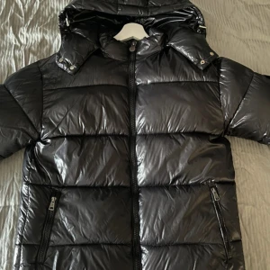 Svart Moncler pufferjacka med huva - Snygg svart pufferjacka från Moncler med glansig finish och huva. Jackan har dragkedja framtill, två fickor med dragkedja och Moncler-logga på ärmen. Insidan har ett coolt serietidningsmönster. Perfekt för kalla dagar när du vill hålla stilen.