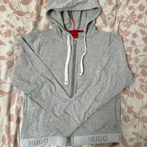 Grå hoodie från Hugo med dragkedja - Snygg ljusgrå hoodie från Hugo med hel dragkedja, vita snören och huva. Tröjan har ribbade muddar och Hugo-logga längs nederkanten. Perfekt för en chill och stilren look. Oanvänd