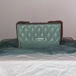 Quiltad grön axelväska med kedja - Snygg quiltad axelväska i ljusgrönt skinnimitation med silverfärgad kedjerem. Väskan har ett stilrent rutmönster och en minimalistisk metall-detalj framtill. Perfekt storlek för det viktigaste och ger en trendig touch till din outfit.