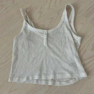 Vit ribbad linnetopp med knappar - Säljer en fräsch vit ribbad linnetopp med croppad passform och smala axelband. Toppen har tre dekorativa knappar framtill och är tillverkad i mjuk bomull. Perfekt för varma dagar och enkel att matcha med jeans eller shorts.