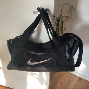 Svart träningsväska från Nike - Stor svart träningsväska från Nike med klassisk vit swoosh-logga på sidan. Väskan har långa axelband, dragkedja upptill och flera fack för smart förvaring. Tillverkad i slitstark polyester och perfekt för gymmet eller sportaktiviteter.