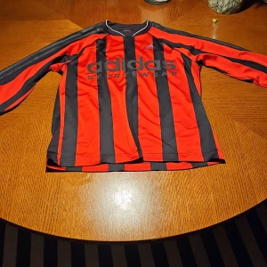 Randig långärmad Adidas fotbollströja - Snygg långärmad fotbollströja från Adidas med klassiska svarta och röda vertikala ränder. Stor Adidas Sportswear-logga på bröstet och tre svarta ränder längs ärmarna. Tillverkad i lätt och ventilerande polyester, perfekt för träning eller match. Tvättlapp bortklippt