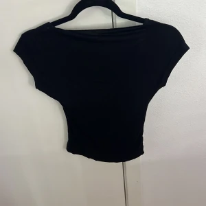 Svart croppad topp med öppen rygg - Säljer en svart croppad topp med korta ärmar och snyggt öppen rygg. Toppen har en enkel, stilren design och är tillverkad i mjukt, stretchigt material som sitter skönt mot kroppen. Perfekt för dig som gillar minimalistisk stil och vill ha något som sticker ut.