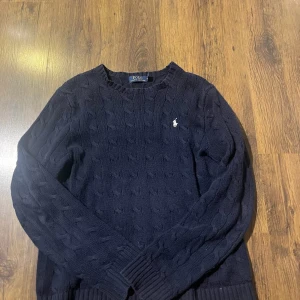 Mörkblå stickad tröja Polo Ralph Lauren - Snygg mörkblå stickad tröja från Polo Ralph Lauren med klassiskt kabelstickat mönster och vit broderad logga på bröstet. Rund hals och långa ärmar med ribbade muddar. Perfekt för dig som gillar stilrena och tidlösa plagg.