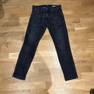 Replay Anbass svarta jeans - Svarta Replay Anbass jeans med snygg tvätt och klassiska fem fickor. Jeansen har smal passform, raka ben och diskreta slitningar. Märkespatch bak i midjan och detaljerade bakfickor. Perfekta för dig som gillar stilrena och moderna jeans.