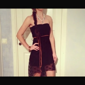 Svart spetsklänning med bälte - Svart axelbandslös asymmetrisk klänning med transparent spets och blommigt mönster. Bältet medföljer. Perfekt för dig som gillar en elegant och unik stil med snygga detaljer. Köpt i en butik i Stockholm, men kommer inte ihåg vilken, då det var så länge sen. Men plagget är i perfekt skick fortfarande. 