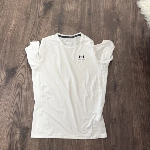 Vit kompressionst-shirt från Under Armour - Säljer en vit kompressionst-shirt från Under Armour HeatGear i storlek 2XL. T-shirten har korta ärmar, rund halsringning och en tight passform. Perfekt för träning med sitt lätta och stretchiga syntetmaterial. Diskret logga på bröstet.