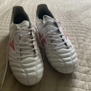 Vita MIZUNO Morelia Neo IV Pro AG Charge - Säljer ett par vita Mizuno fotbollsskor med röda detaljer på sidorna. Skorna har snörning och är tillverkade i skinn för bästa känsla på planen. Endast använda 3-4 gånger men säljer då de var för stora. Nypris: 1000kr