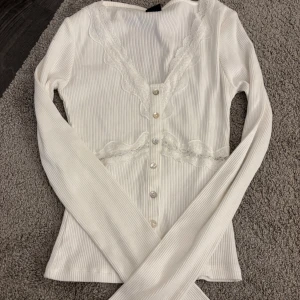 Vit ribbad blus med spets från Gina Tricot - Supersöt vit ribbad blus från Gina Tricot i storlek S. Den har långa ärmar, v-ringning med spetsdetaljer och knappar framtill. Spetsen går även runt midjan och ger en extra feminin touch. Perfekt för dig som gillar romantisk stil!