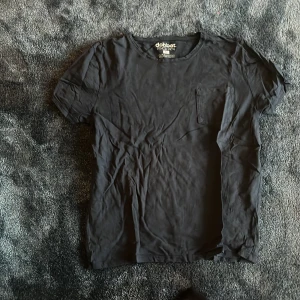 Svart t-shirt från Dobber, storlek M - Svart t-shirt från Dobber med rund hals och en liten bröstficka. Modellen är klassisk och har korta ärmar. Materialet är mjuk bomull som känns skönt mot huden. Perfekt för dig som gillar enkel och stilren design.