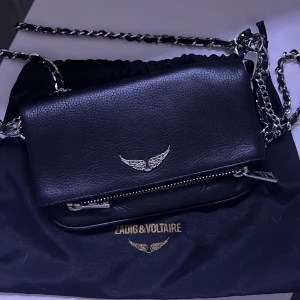 Svart axelväska från Zadig & Voltaire - Snygg svart axelväska i skinn från Zadig & Voltaire med silverfärgad kedjerem och ikonisk vinge i metall framtill. Väskan har dragkedja och stilren design, perfekt för dig som gillar edgy detaljer och vill ha något som sticker ut.