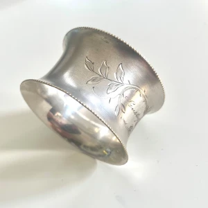 Antik Servettring- WMFM- Silverpläterad- Graverad med namn - Här är en antik, vacker servettring i silverpläterat material från den välkända tyska tillverkaren WMFM (Württembergische Metallwarenfabrik). Designen är tidstypisk för slutet av 1800-talet och tidigt 1900-tal, Art Nouveau. Den är i mycket gott skick med minimalt slitage för sin ålder. En vacker och historiskt intressant detalj för samlaren eller varför inte som gåva till någon med namnet Gustaf? Mått: H 3 cm Omkrets 5 cm Vikt 30,65 gram
