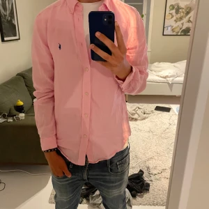 Rosa linneskjorta från Ralph Lauren - Säljer en snygg rosa skjorta från Ralph Lauren i linne med klassisk krage och blå broderad logga på bröstet. Skjortan har lång ärm, knappar framtill och en avslappnad passform. Perfekt för dig som vill ha en fräsch och stilren look. Det är storlek xl i barn men passar som M. Jag är 181.
