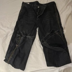 Svarta jeans med dragkedjor från Zara - Unika svarta jeans från Zara med råa, slitna detaljer och stora dragkedjor längs benen. Jeansen har en loose fit och är tillverkade i kraftig denim. Perfekta för dig som vill sticka ut med en edgy streetstyle-look.
