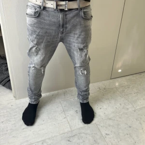 Grå slitna jeans med hål - Säljer ett par grå jeans med slitningar och hål på både fram- och baksida. Jeansen har en tapered passform och normal midja. De är tillverkade i denim och har en cool, streetig vibe med sina trasiga detaljer.